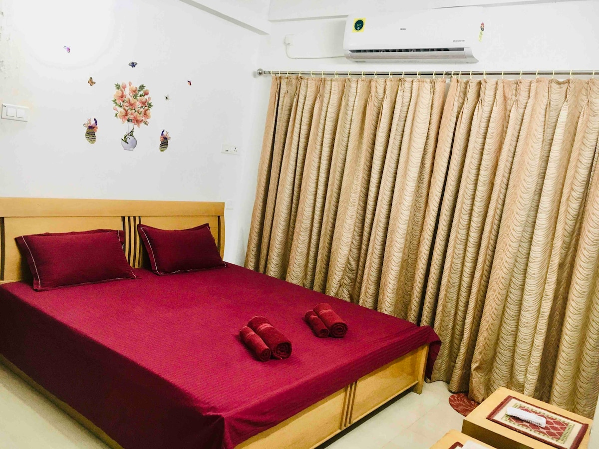 A well-lit private room features a spacious bed adorned with deep red bedding and matching rolled towels. Elegant patterned curtains frame a window, while floral wall decor adds a touch of warmth. A small table is positioned nearby, creating a cozy sleeping environment.
