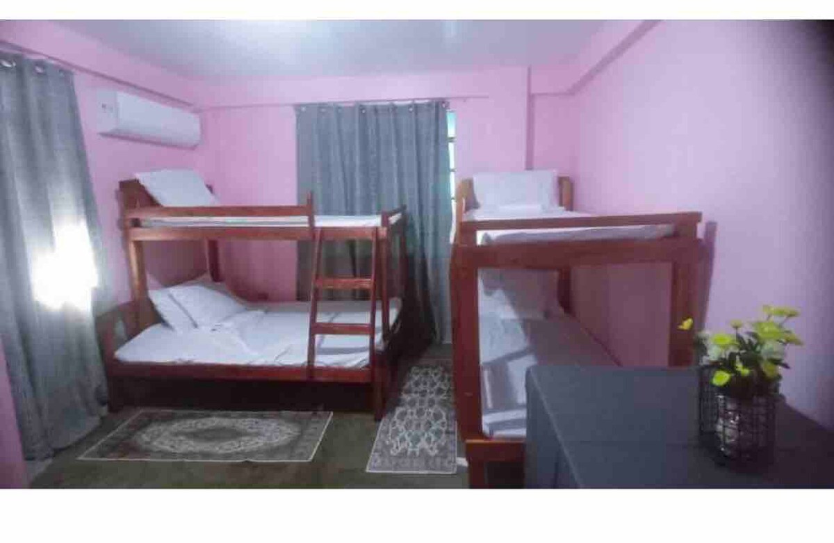 The room features two wooden bunk beds arranged across from each other, providing ample sleeping space. Soft bedding is neatly laid out on each level, with curtains hanging on the window. A small table with a plant adds a touch of comfort to the corner.