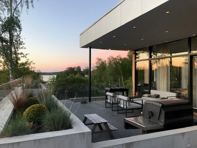 An elegant outdoor terrace is featured, showcasing a spacious seating area with modern furniture. A grill is positioned nearby, set against a backdrop of lush greenery. The soft hues of sunset reflect on the water, creating a serene atmosphere.