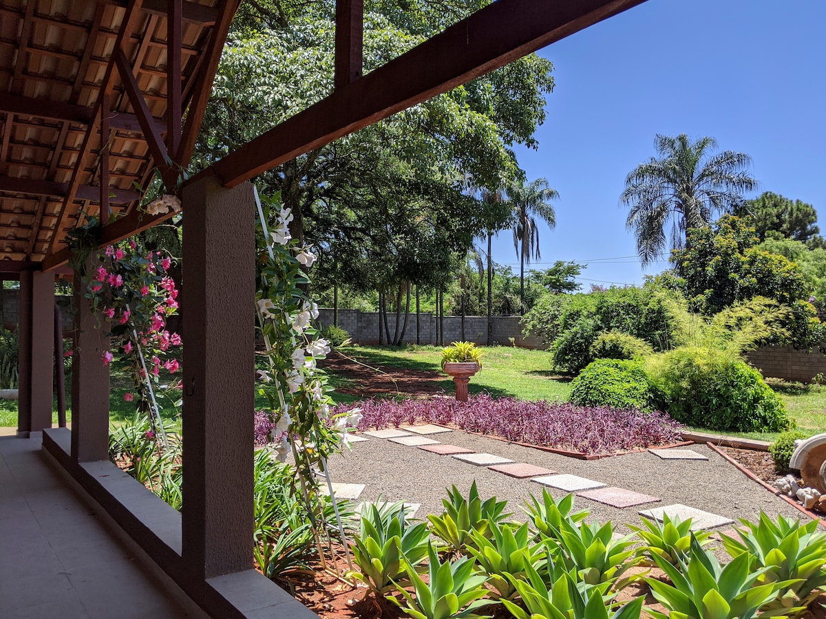 A spacious outdoor area is framed by a covered porch, showcasing a lush garden filled with colorful foliage. Flowering plants line the pathway, leading to a central planter. Tall trees provide shade, creating a peaceful environment ideal for relaxation.