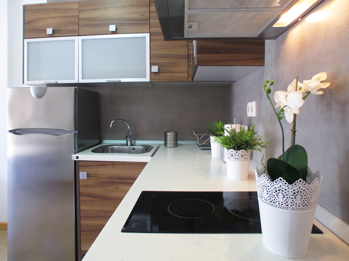 A modern kitchen is shown, featuring sleek wooden cabinets and a light-colored countertop. Stainless steel appliances, including a refrigerator, are visible alongside a sink. Potted plants are placed decoratively, adding a touch of greenery to the space.