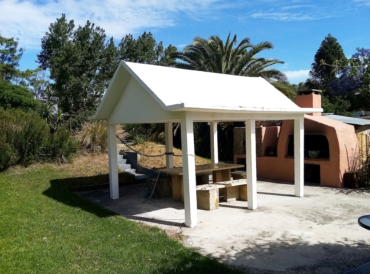 An outdoor area features a covered barbecue structure with a table and benches. Surrounding greenery includes palm trees and shrubs, enhancing the peaceful setting. A stone fireplace is visible in the background, contributing to the space's functionality for gatherings.