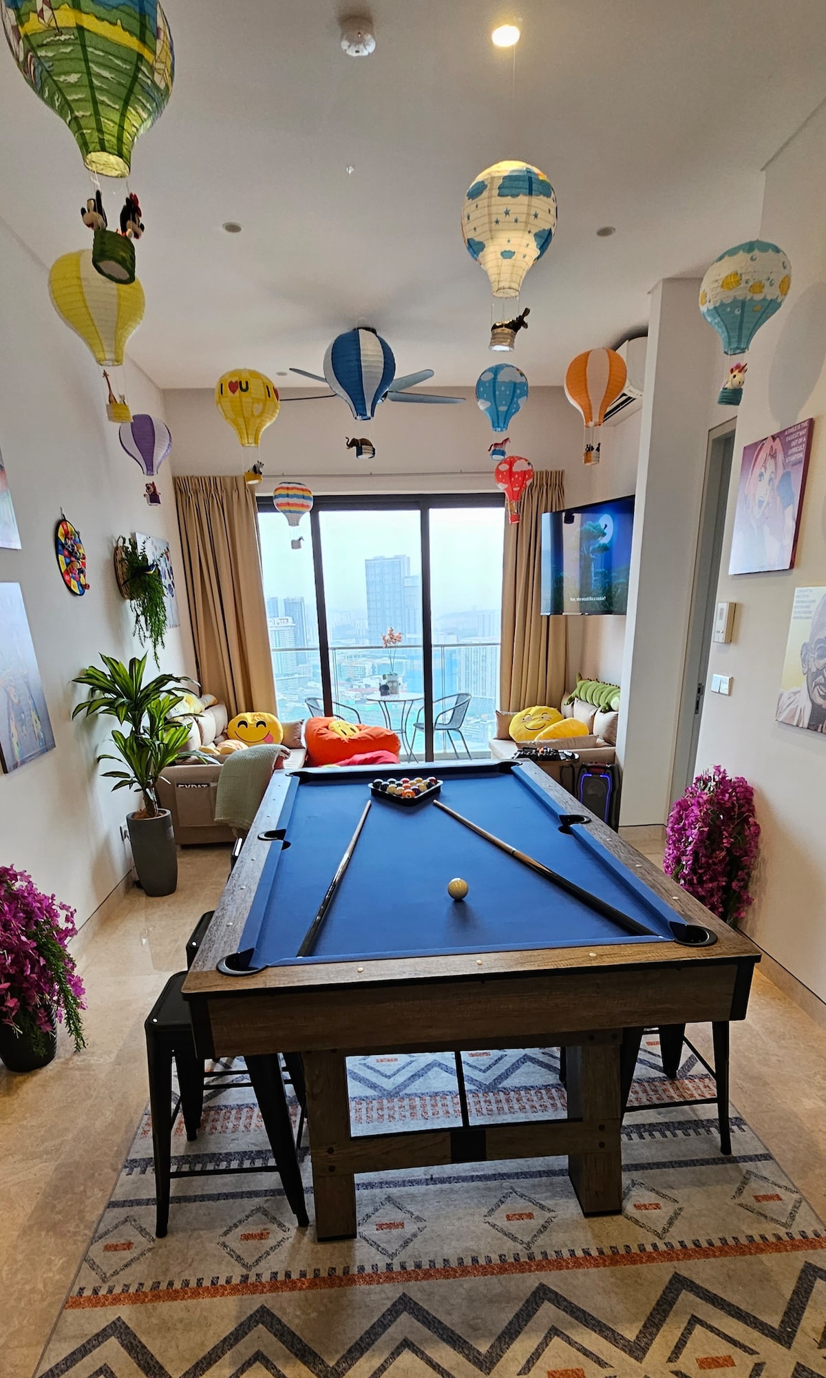 A vibrant game room features a spacious pool table centered beneath colorful hot air balloon decorations. Large windows provide city views, while potted plants and decorative elements add a playful touch to the space.