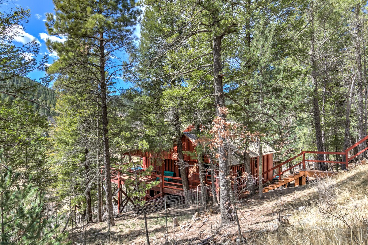 A rustic cabin, nestled among tall pine trees, features a wraparound deck and can be accessed via a gently sloping path. The natural setting is complemented by vibrant blue skies, creating a serene atmosphere in the woods.