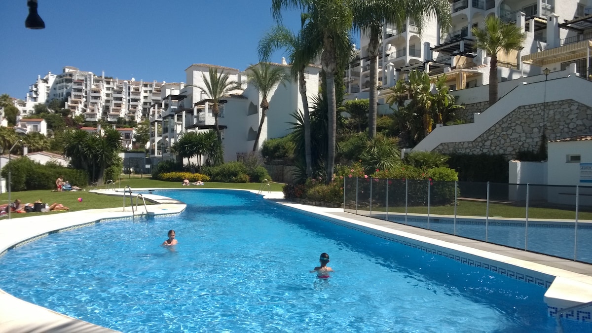 A large swimming pool is surrounded by palm trees and landscaped gardens. Sunbathers are seen relaxing on the poolside. In the background, white buildings are positioned on a hillside, contributing to the community's vibrant atmosphere.