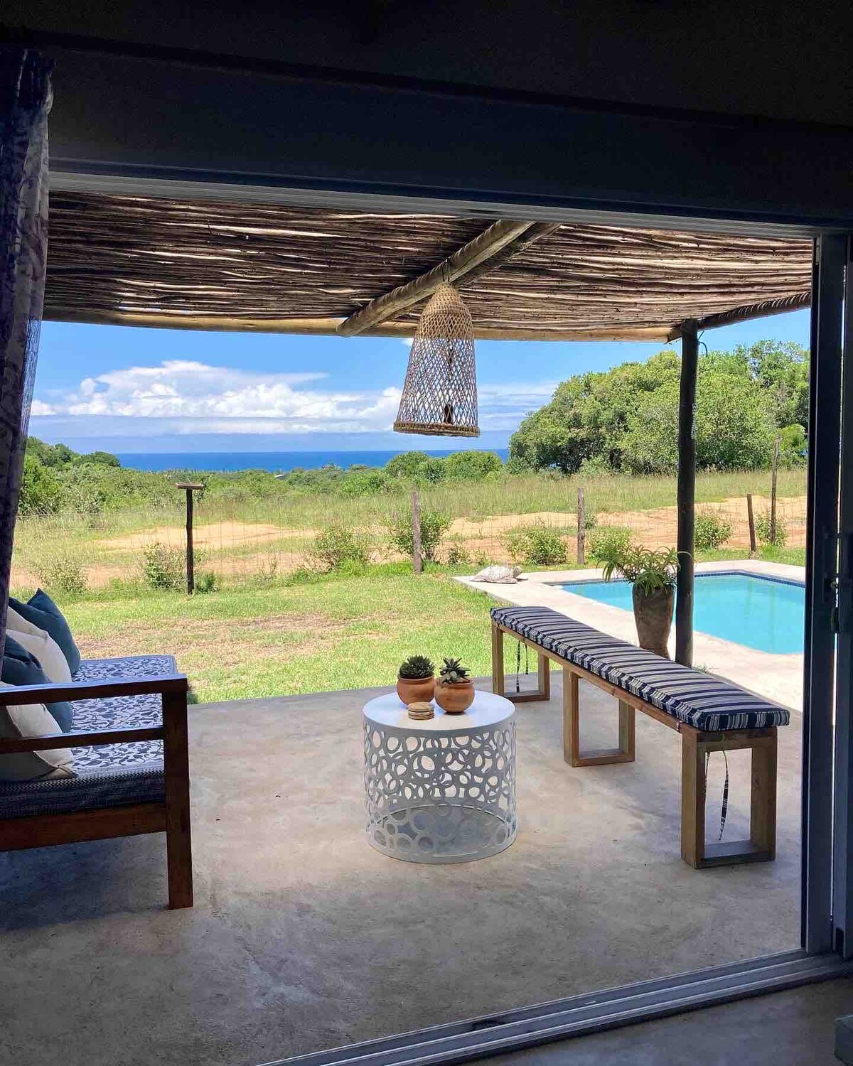 A serene outdoor seating area is visible, featuring a wooden bench with patterned cushions and a round table with potted plants. The space overlooks a lush landscape leading to a distant view of the ocean. A refreshing pool is nearby, complemented by natural light from the open setting.