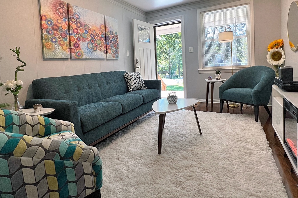 A spacious living room is furnished with a blue sofa, a patterned armchair, and a minimalist coffee table. A large window allows natural light to brighten the space, which features a soft rug underfoot and colorful wall art enhancing the decor.