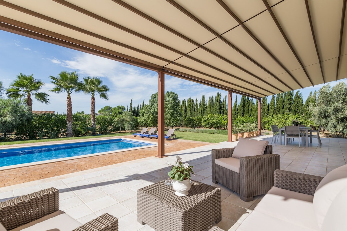 A shaded outdoor terrace features comfortable seating arrangements and a coffee table. The large swimming pool is visible in the distance, surrounded by vibrant greenery and palm trees. A dining area can be seen off to one side, providing space for outdoor meals.