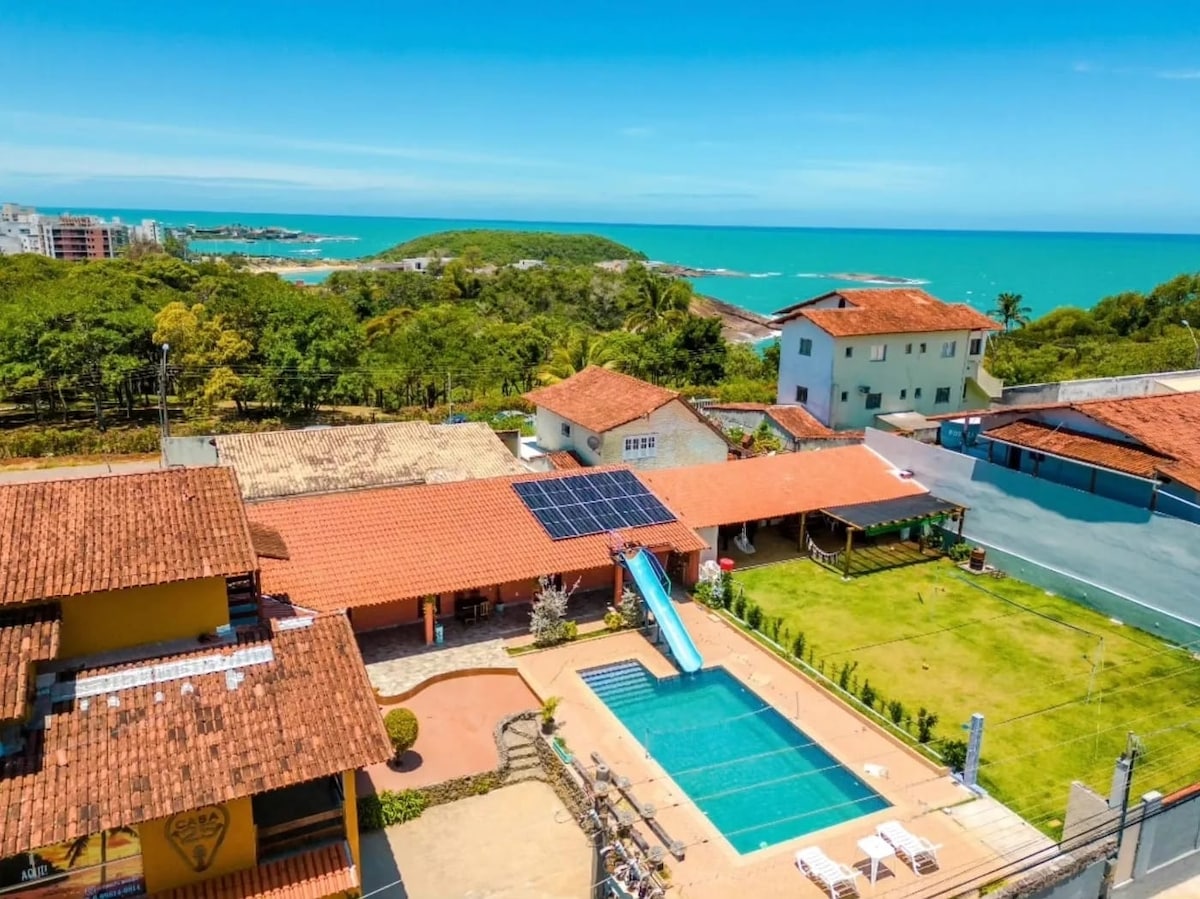 The image captures an aerial view of a vibrant property, featuring a clear blue pool in the foreground, surrounded by well-maintained grass and lounge chairs. A large waterslide is visible, along with neighboring buildings and a glimpse of the ocean in the background under a bright blue sky.