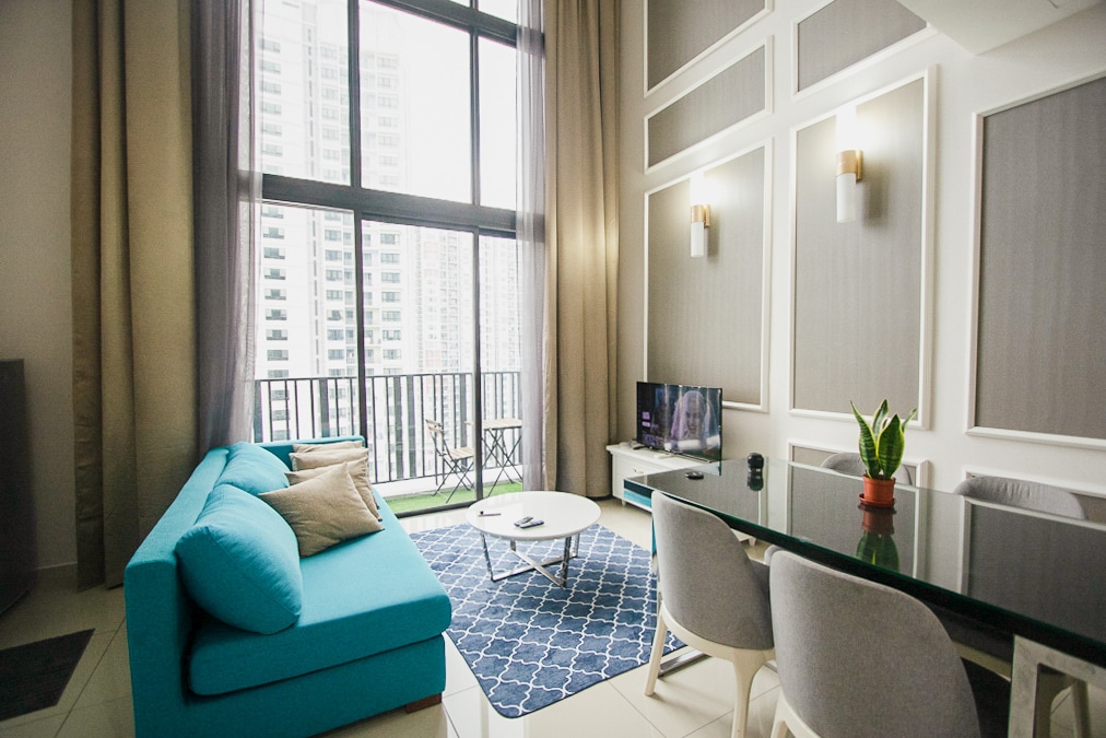 A contemporary living area features a blue sofa with accent cushions near a glass coffee table. Soft curtains frame large windows that offer views of the city skyline. A small dining table with light-colored chairs is positioned adjacent to the sofa, complemented by a potted plant.