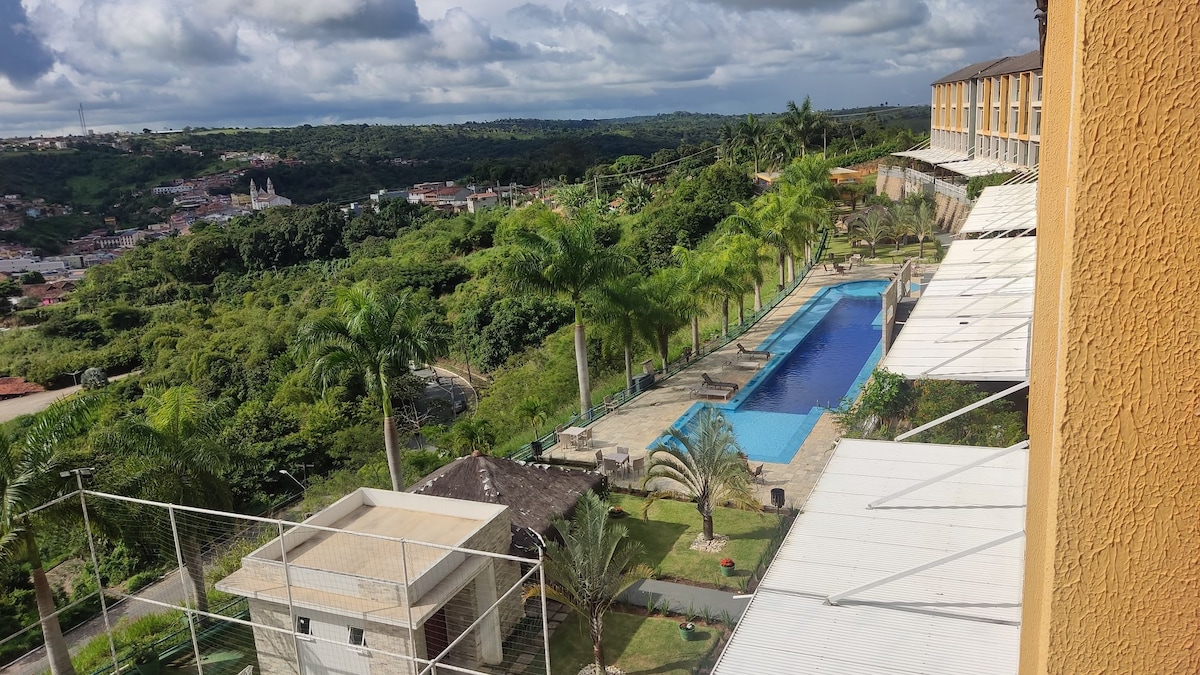 A panoramic view showcases a serene landscape featuring a long swimming pool surrounded by palm trees. The green hills in the distance can be seen, alongside the nearby structures, offering a sense of tranquility in this elevated setting.