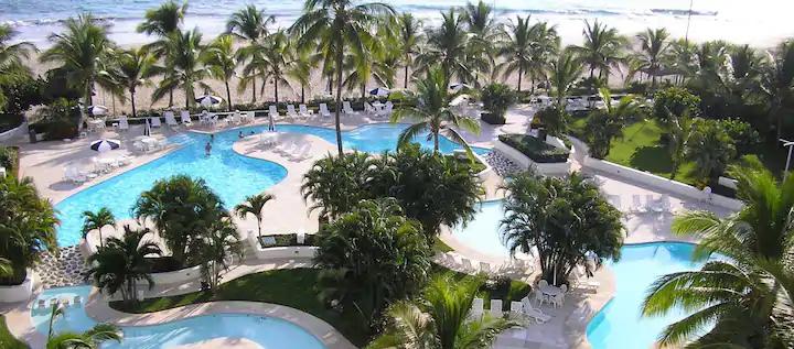 A view of the expansive outdoor pool area is featured, surrounded by lush palm trees. Multiple swimming pools are arranged together, with sun loungers positioned around them. The beach and ocean can be seen in the distance, enhancing the tranquil atmosphere.