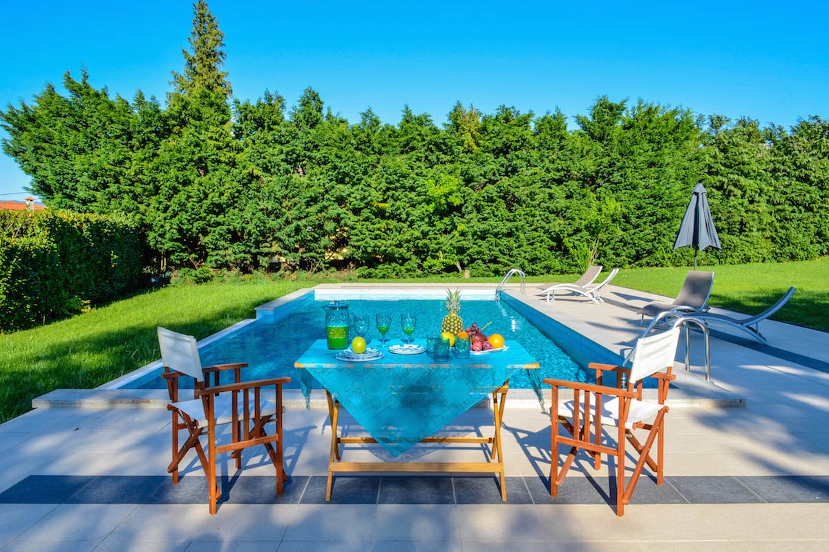 A serene swimming pool is surrounded by well-maintained greenery. A dining table is set with various fruits and drinks under a bright blue sky. Two lounge chairs and an umbrella are positioned nearby, creating a welcoming outdoor space for relaxation.
