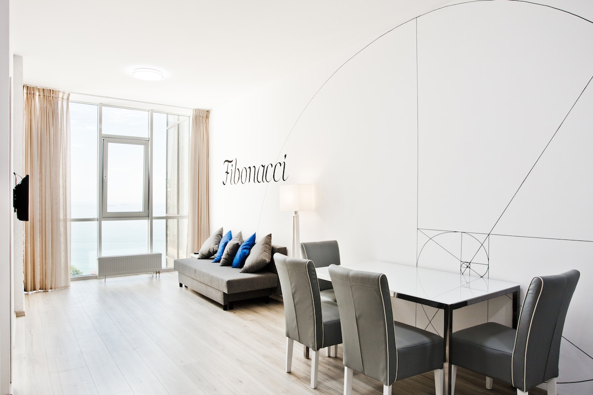 A spacious living area features large windows with a direct view of the sea. A light-colored sofa is adorned with blue accent pillows, and a dining table with grey chairs is positioned nearby. The wall is decorated with a design inspired by Fibonacci numbers.