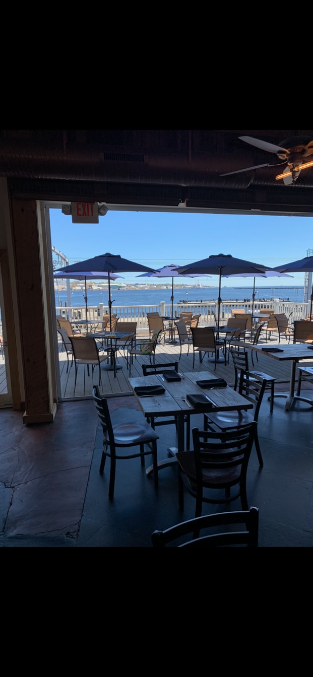 Outdoor seating is arranged on a deck with several tables and chairs shaded by large umbrellas. The view beyond showcases the waterfront and distant structures against a clear blue sky.