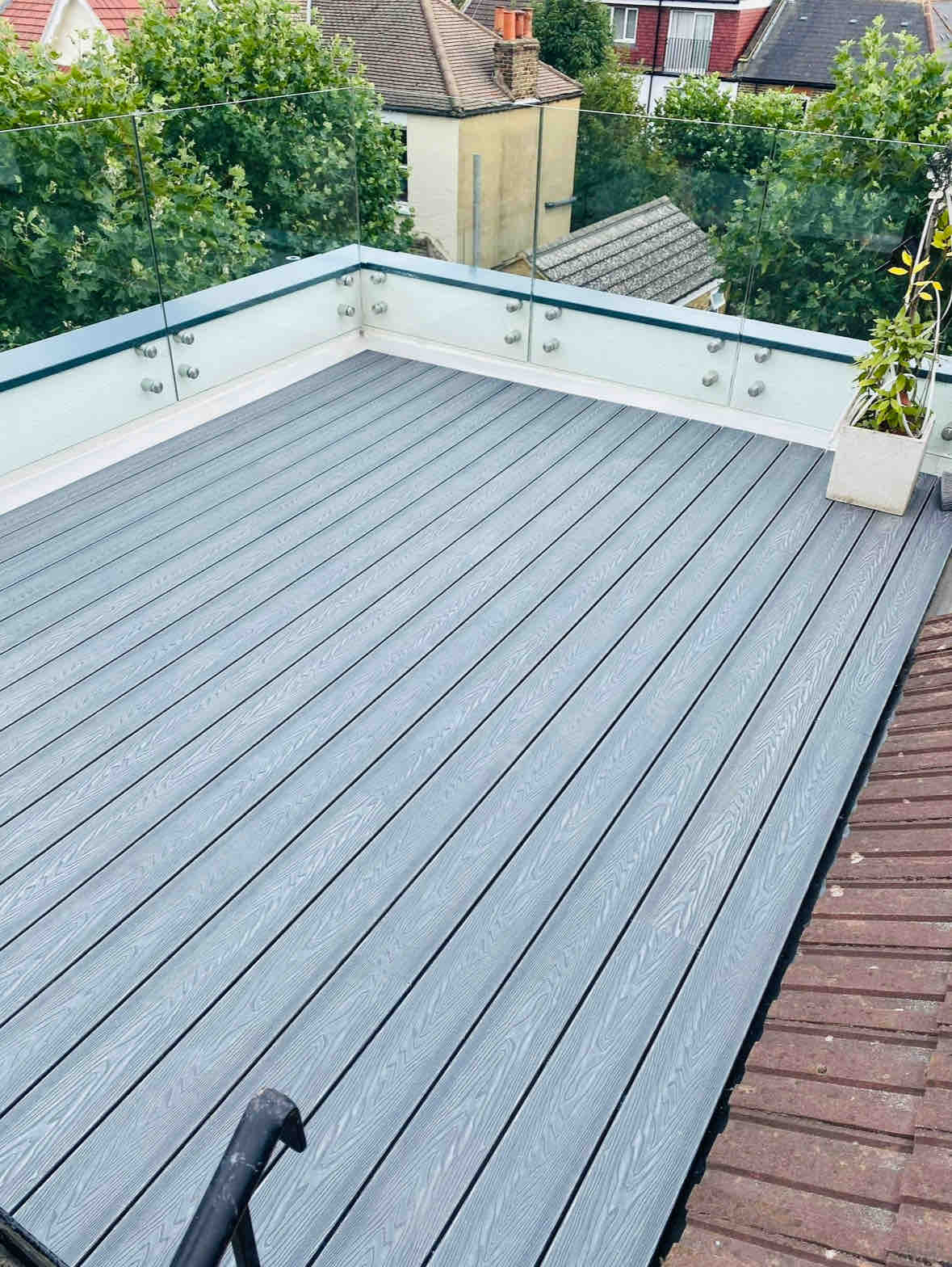 The private roof terrace features a wooden deck with a grey finish, surrounded by clear glass panels. Potted greenery adds a touch of nature, while the view showcases nearby rooftops and treetops, creating an inviting outdoor space for relaxation.