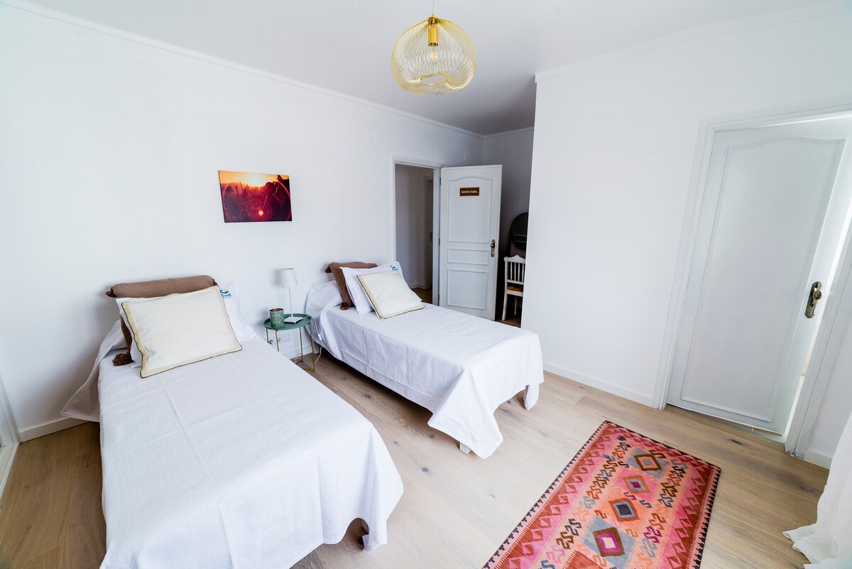 A bright room is furnished with two separate beds, each dressed in white linens, and accented by light-colored pillows. A colorful rug adds warmth to the light wood floor. Natural light brightens the space, complemented by a light fixture overhead and a framed print on the wall.