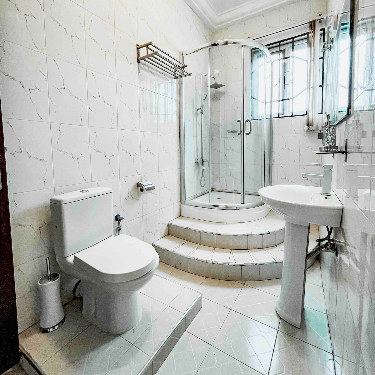 A modern bathroom features a glass shower stall with a curved design and tiled steps leading up to it. A white toilet and pedestal sink are positioned on opposite sides, with sleek fixtures and a window allowing natural light to fill the space.