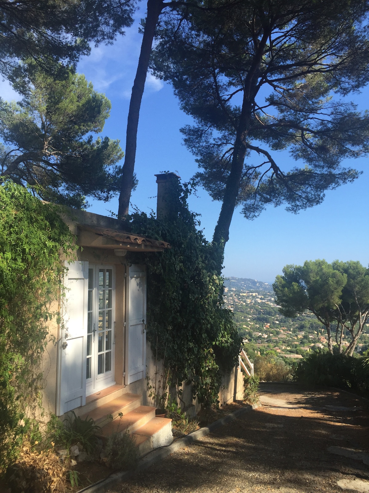 A charming entrance is framed by lush greenery and tall pine trees. The light-colored building features double white doors set within a stone wall, leading to the serene surroundings. The sky above is clear with gentle blue hues, offering a glimpse of the valley below.