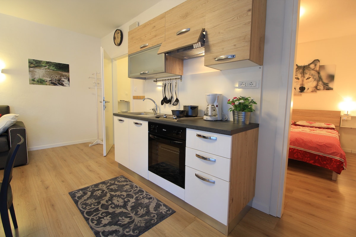 A compact kitchenette is visible, featuring modern cabinetry and essential appliances including a microwave, toaster, and coffee maker. Hanging utensils and a small potted plant add to the functional space. Natural light illuminates the area, connecting it to a nearby living space with cozy seating.