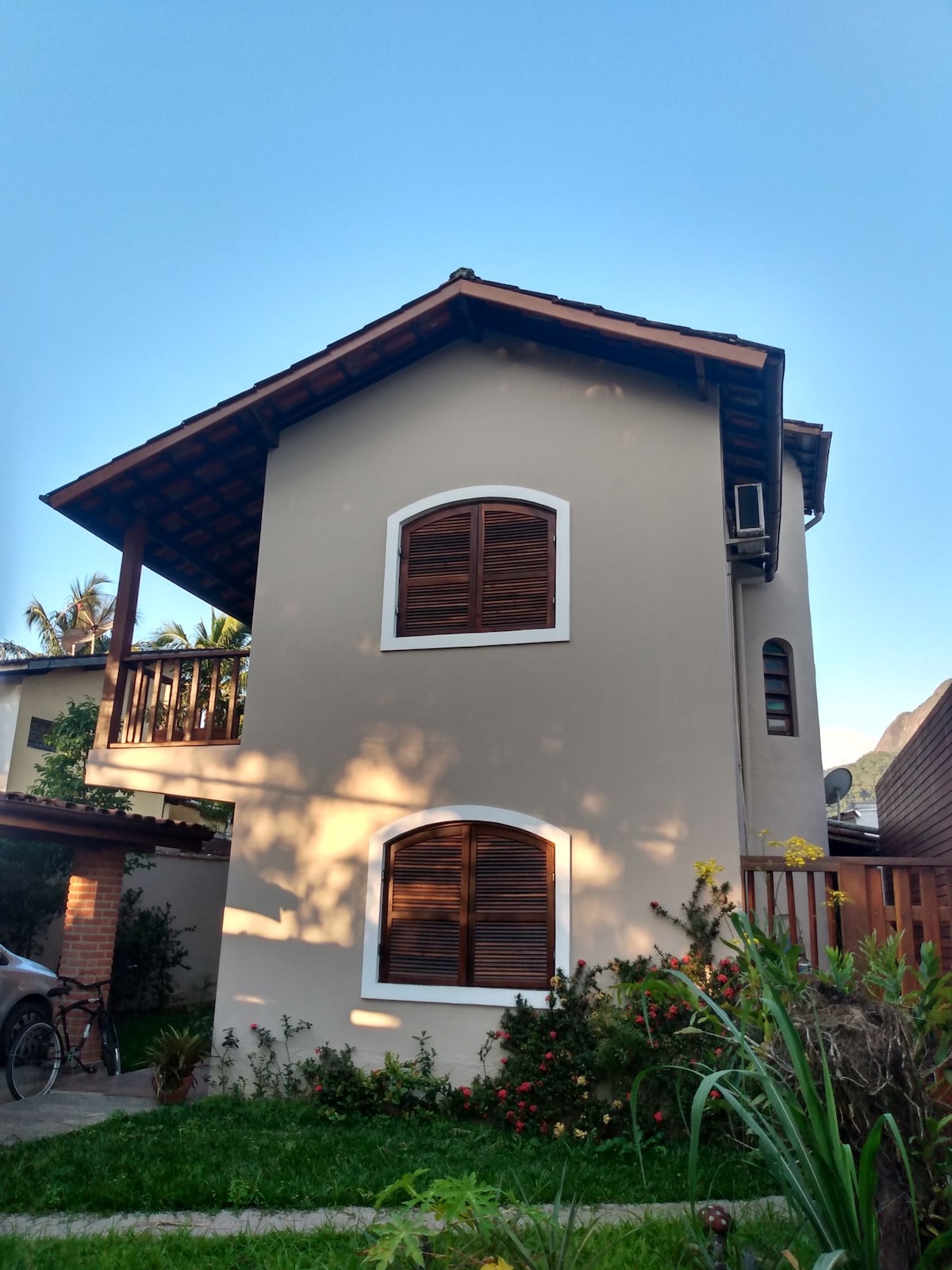 A two-story house is displayed, featuring a neutral-toned exterior with wooden shutters on the windows. A covered balcony is visible on the upper level, surrounded by greenery and flowers in the foreground. The area appears well-maintained and inviting.