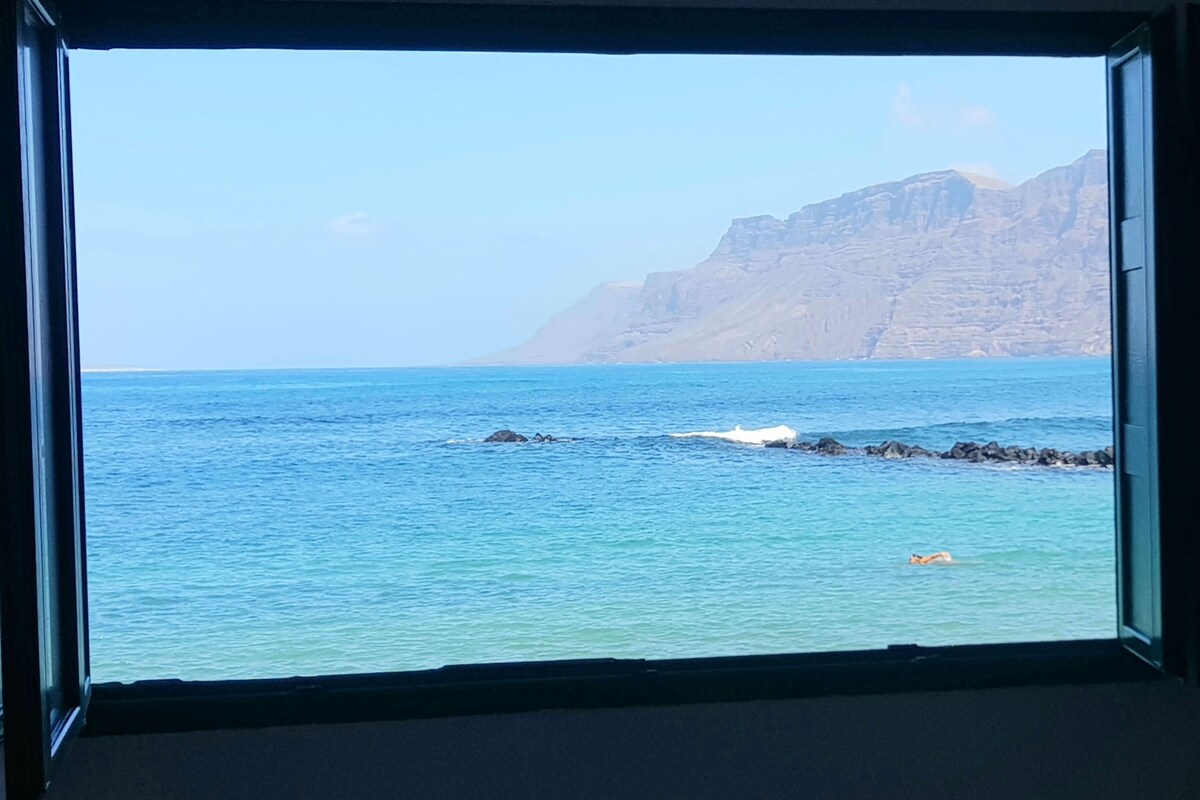 A large window frames a panoramic view of the ocean, showcasing clear blue waters meeting rocky shores and a distant mountain range. The tranquil scene is highlighted by gentle waves and a solitary figure enjoying the water.
