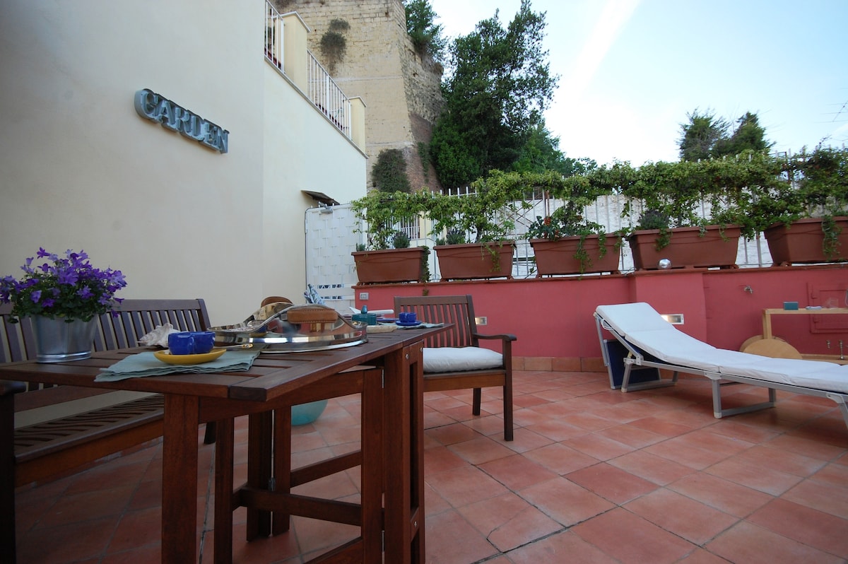 An outdoor terrace space features a wooden dining table and several comfortable seating arrangements. Potted plants line the wall, providing greenery. A sun lounger is positioned for relaxation, and the area is adorned with colorful accents, creating an inviting environment for outdoor enjoyment.