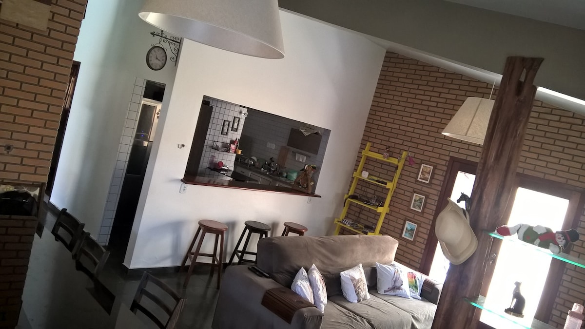 The living area showcases a spacious layout with large windows allowing natural light. A comfortable seating arrangement is present, accompanied by two wooden stools near a bar counter. Decor elements include rustic brick walls, highlighting the contemporary design.