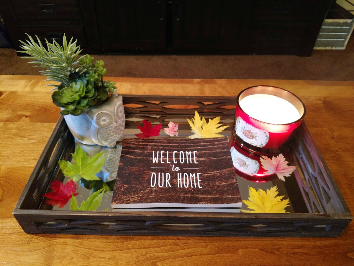 A decorative tray is arranged with a potted succulent and a lit candle, surrounded by colorful autumn leaves. A welcoming note resides in the center, adding a personal touch to the inviting display.