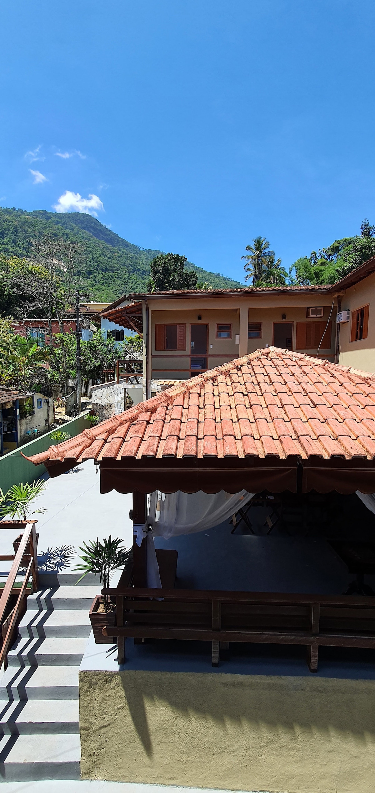 An outdoor view captures a charming structure with a red-tiled roof, surrounded by lush foliage and distant mountains. The scene includes a welcoming terrace area adorned with drapes, providing a shaded space for relaxation. Blue skies enhance the serene atmosphere.
