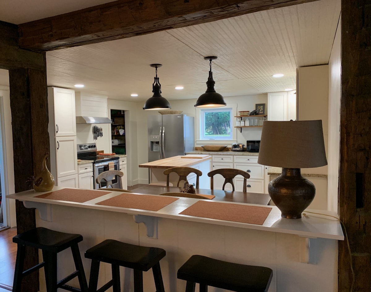 The kitchen space features modern appliances and a central island with seating. Under-cabinet lighting illuminates the work area. Black bar stools are positioned at the counter, complemented by decorative lamps and rustic wooden beams that enhance the cozy atmosphere.
