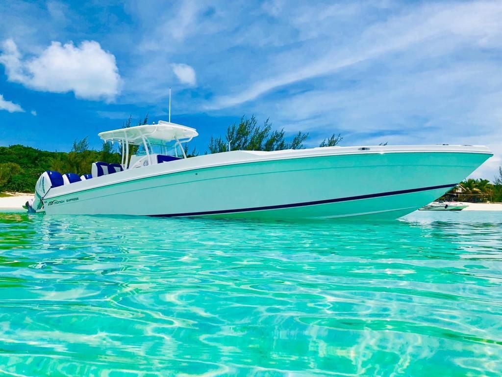 A sleek boat is anchored in clear turquoise waters, surrounded by a pristine beach and lush greenery in the background. The vessel's exterior features a clean white finish with blue accents, providing a modern and inviting appearance.