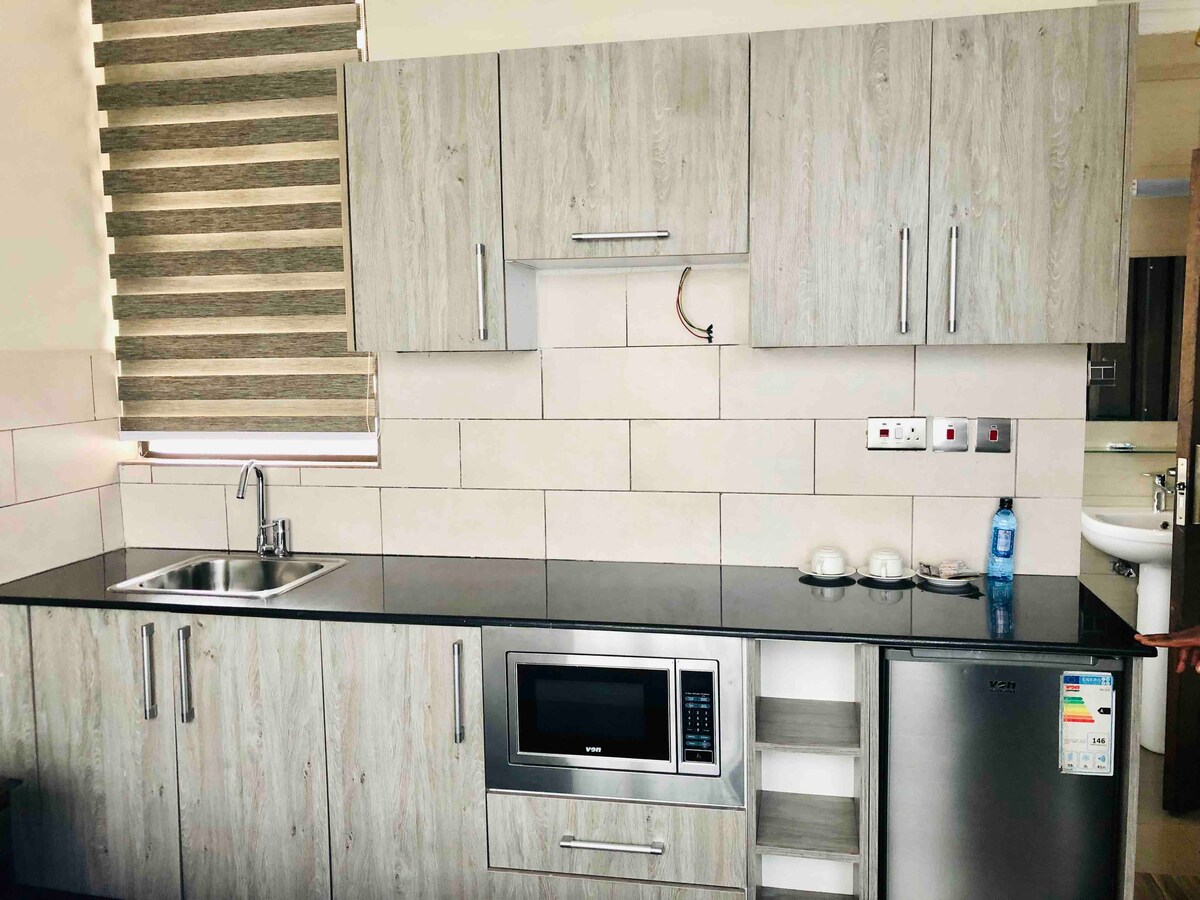 A modern kitchen features light wood cabinetry paired with a dark countertop. Notable appliances include a microwave and a stainless steel refrigerator. A stainless steel sink is positioned under a window adorned with striped blinds, allowing natural light to brighten the space.
