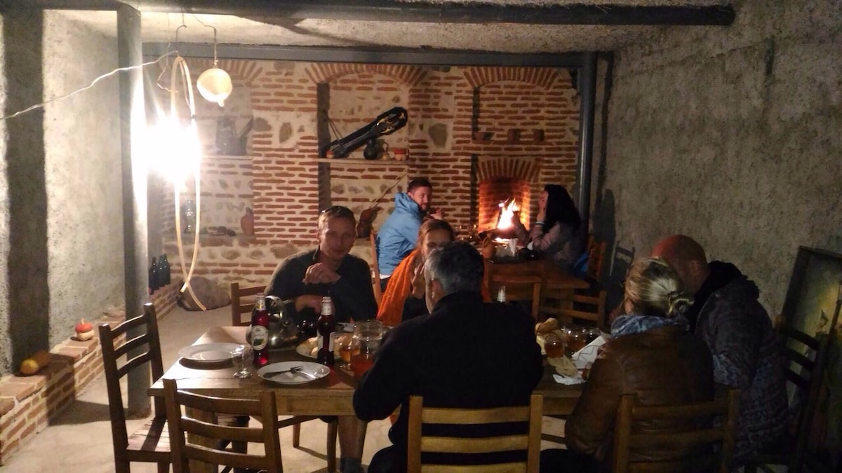 A dining space is captured with a rustic ambiance, featuring wooden tables and chairs arranged around a central area. A warm fireplace adds to the cozy atmosphere. Textured stone walls and brick details create a traditional backdrop for gatherings.