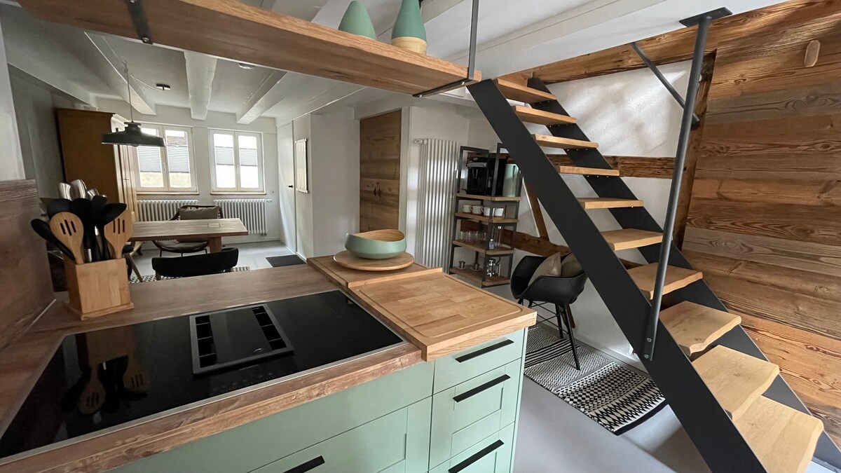 An open kitchen features modern cabinetry in soft green, complemented by a wooden countertop. A cooking island is centrally located, with cooking utensils neatly organized in a wooden holder. In the background, a dining area with a wooden table is visible, and a staircase leads to the upper level.