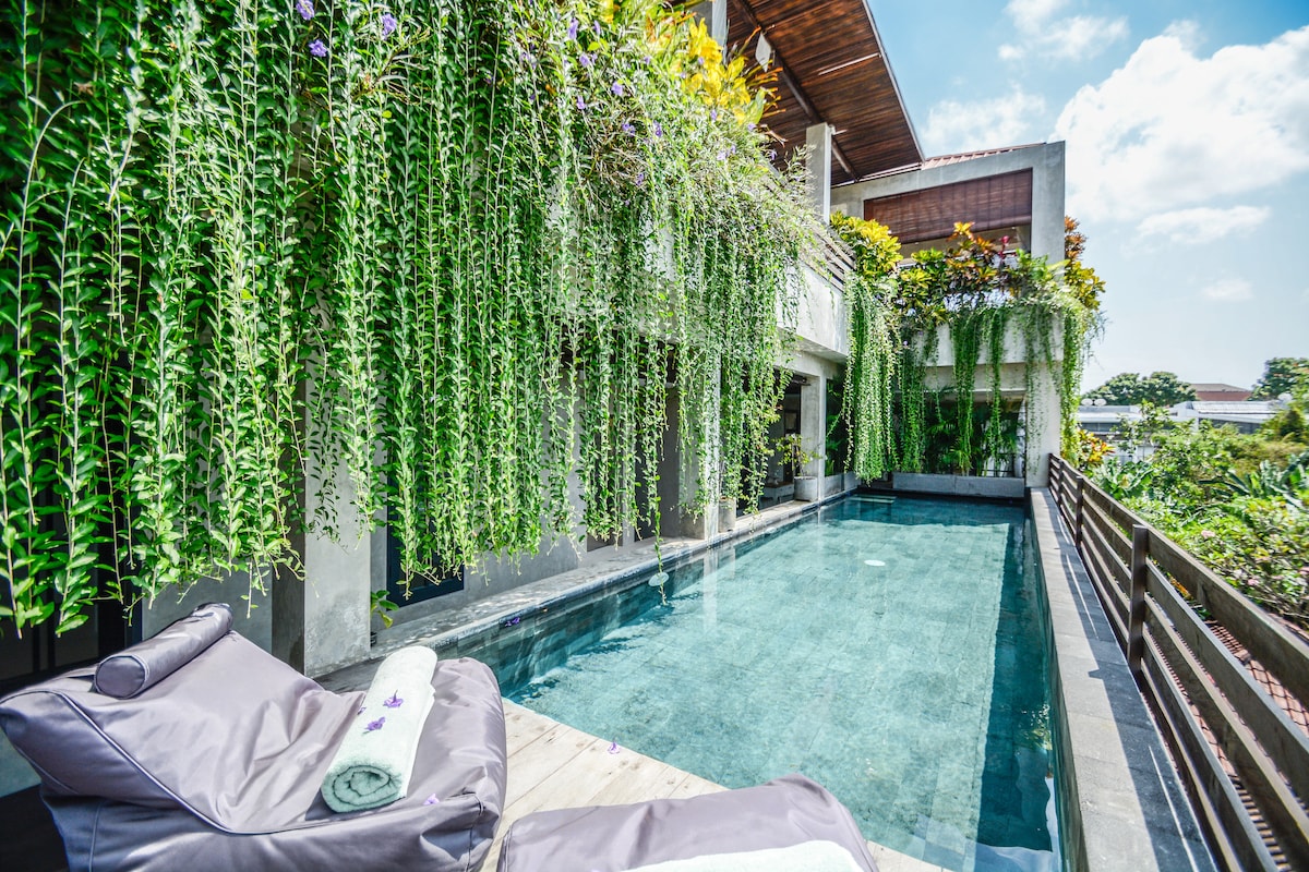 An outdoor area features a serene swimming pool surrounded by lush greenery, with cascading plants hanging above. Comfortable bean bags are positioned nearby, providing a relaxing spot to enjoy the tranquil setting.