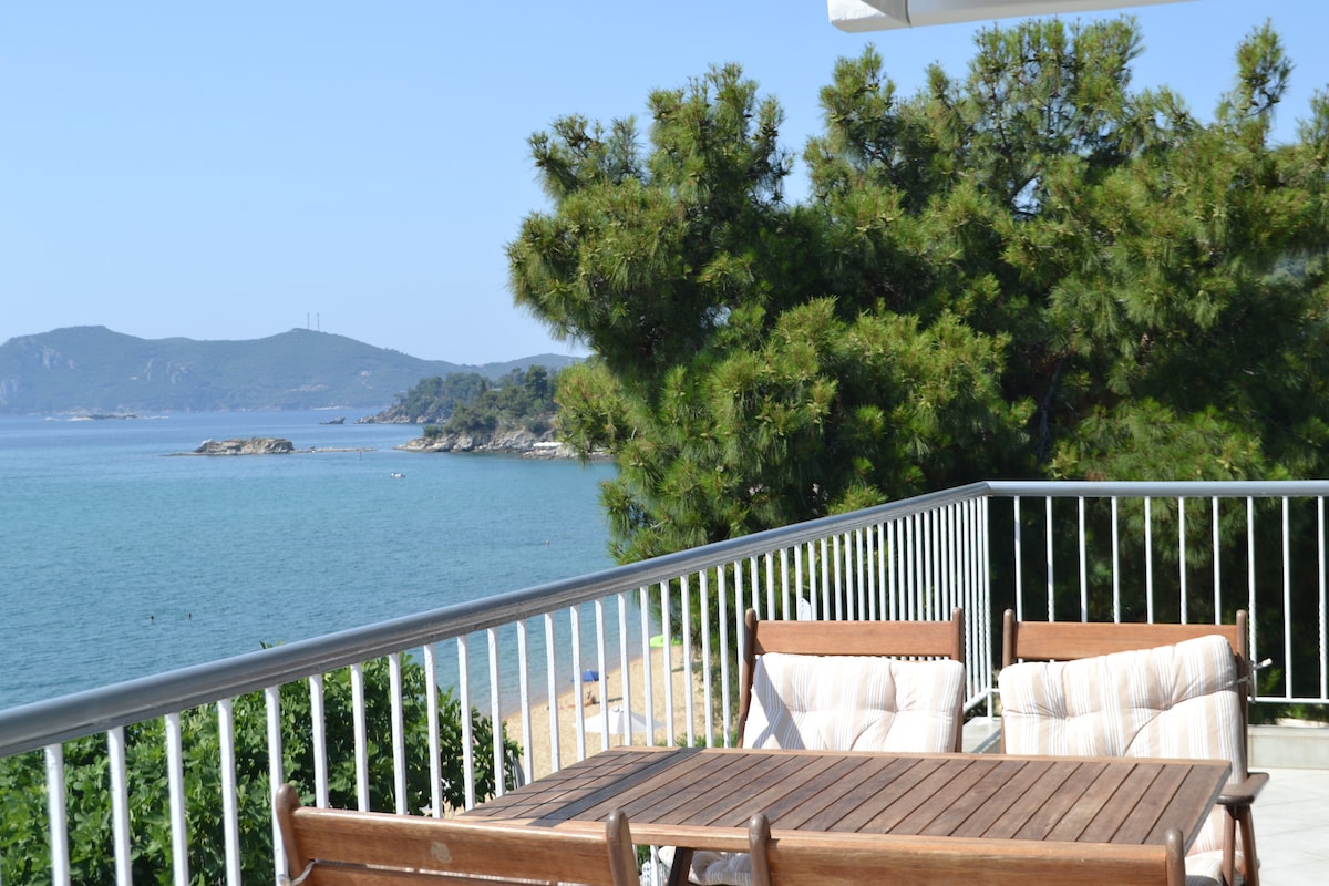 A spacious balcony overlooks the serene sea, with wooden furniture and comfortable cushions arranged for relaxation. Lush greenery borders the view, and distant hills can be seen across the water, creating a tranquil seaside atmosphere.