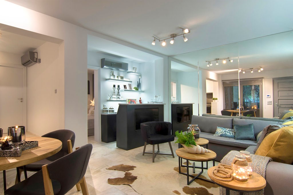 A modern living area is displayed, featuring a sleek bar counter and a cozy seating arrangement. Natural light fills the space, highlighting the neutral color palette and finishes. The dining area is visible with a round table and four chairs, creating a welcoming environment for gatherings.