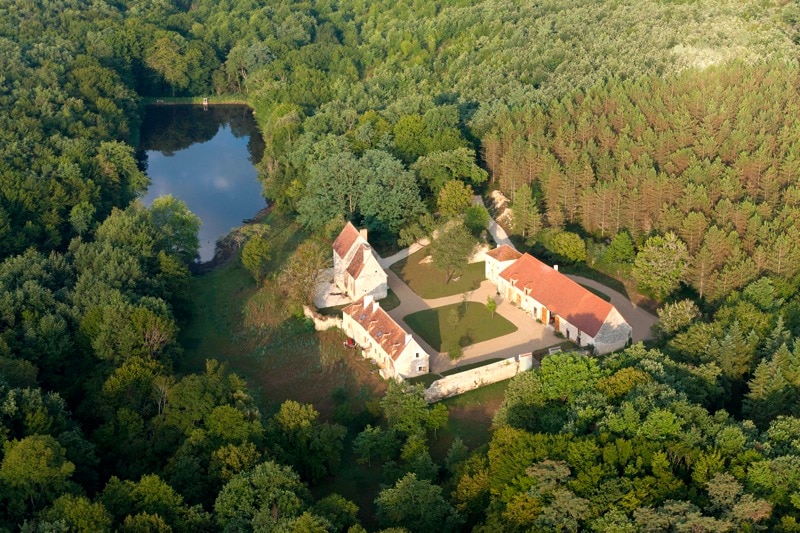 An aerial view captures a charming property nestled within lush greenery. The complex includes several buildings with terracotta roofs and is surrounded by dense forest, along with a glistening pond reflecting the landscape.