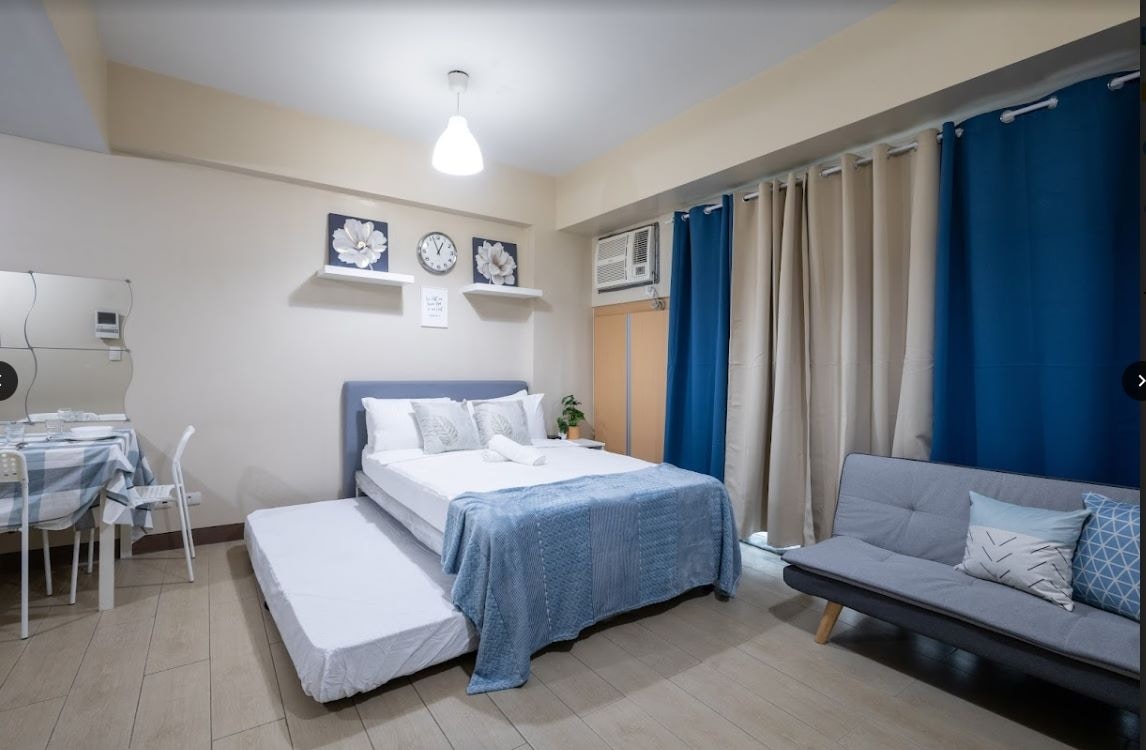 A studio bedroom features a spacious bed with light bedding, complemented by a patterned blue throw. A gray sofa bed with decorative pillows and a small dining area, adorned with a blue and white tablecloth, are also visible. Natural light filters through the blue curtains.