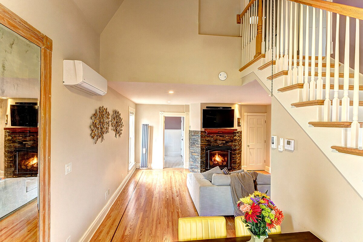 A bright living area features hardwood flooring, a cozy fireplace surrounded by stone, and large windows providing ample natural light. A comfortable sofa with a light throw is positioned near a dining area, where a vase of colorful flowers adds a cheerful touch.