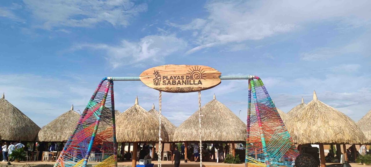 A welcoming entrance to Playas de Sabanilla is marked by a wooden sign featuring the beach's name. Traditional thatched-roof huts, constructed with natural materials, are visible in the background under a clear sky, contributing to a relaxed coastal atmosphere.