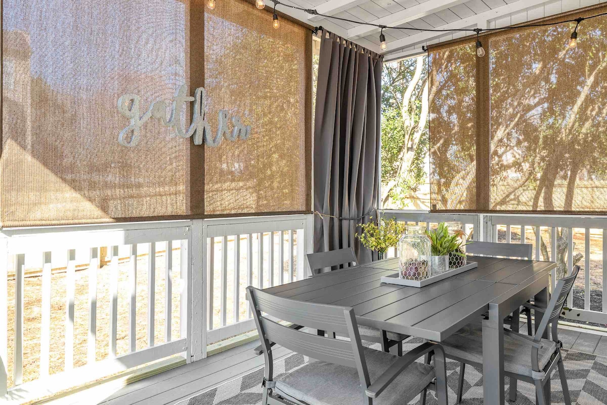 A screened porch features an outdoor dining table surrounded by comfortable chairs. Natural light streams through the screened panels, and walls are adorned with simple decor. The space is illuminated by cafe-style lighting, creating an inviting atmosphere for shared meals.