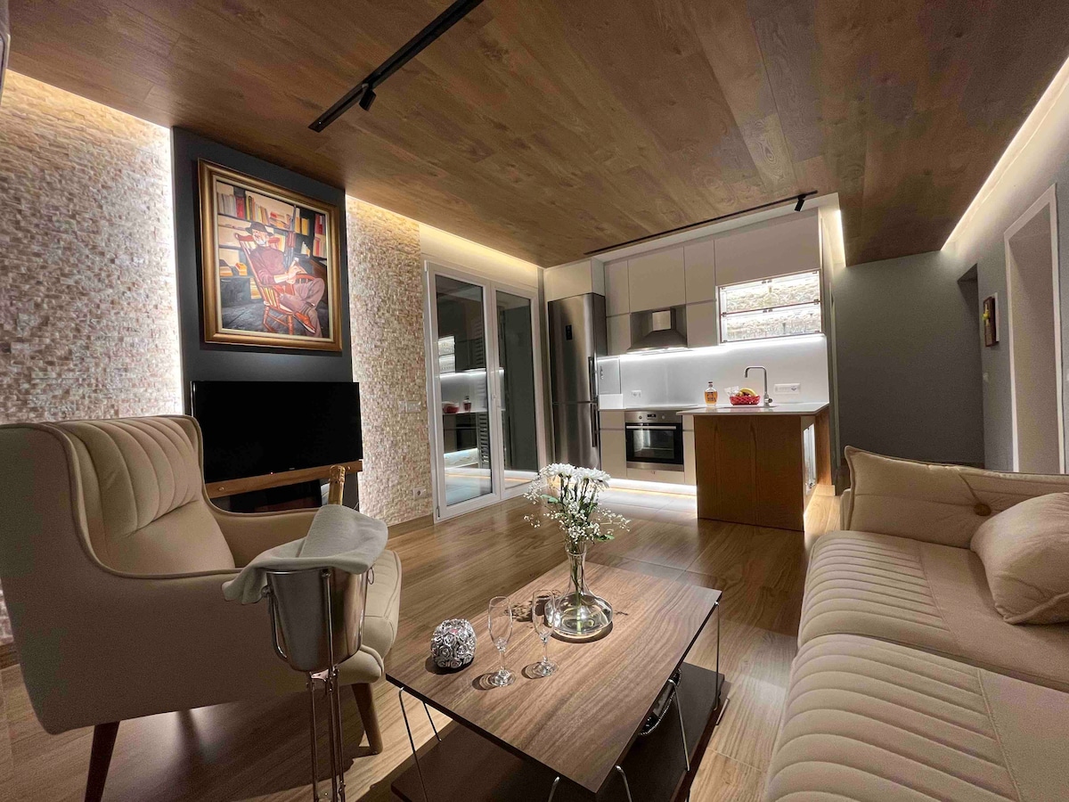 A modern living area is featured, showcasing a beige sofa and a stylish armchair. Soft lighting highlights the textured wall and wooden ceiling. A small coffee table holds glassware, while an art piece adds character to the space. Large glass doors allow natural light to flood in.