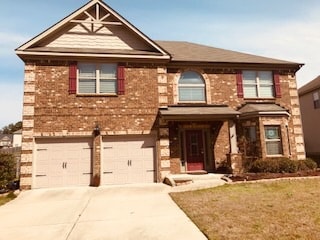 A two-story brick house features a welcoming entrance with a decorative arch above the front door. The exterior showcases shutters on each window. A concrete driveway leads to a double garage with paired doors, bordered by low landscaping.