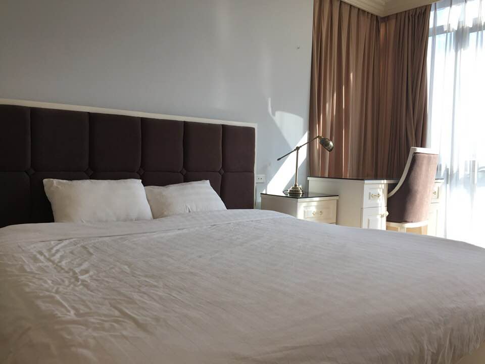 A spacious bed is positioned against a tufted headboard, complemented by soft, natural light filtering through sheer curtains. A tastefully arranged bedside table with a lamp stands nearby, while a stylish chair is visible beside a window, enhancing the room's serene ambiance.