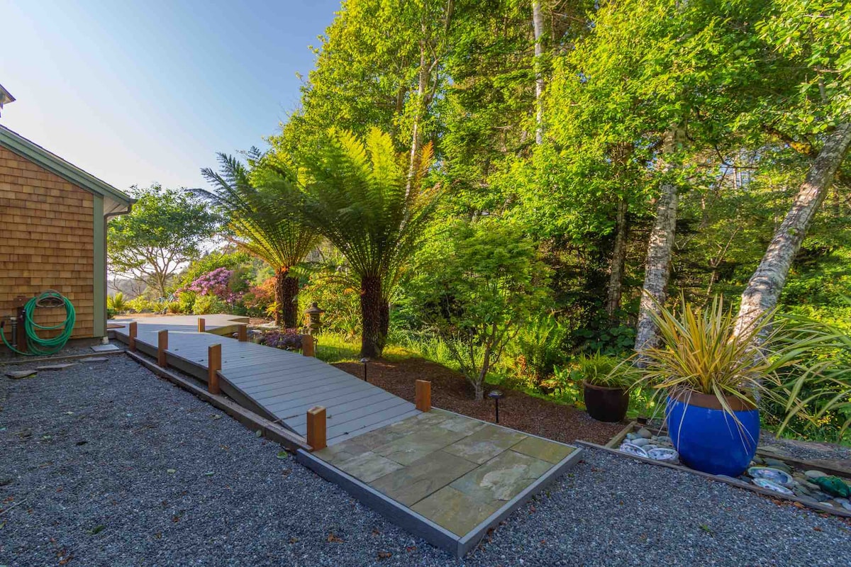 A stone pathway leads through a lush garden, bordered by tall ferns and various trees. Natural sunlight filters through the foliage, creating a serene environment with gravel underfoot. Colorful planters add visual interest along the path.