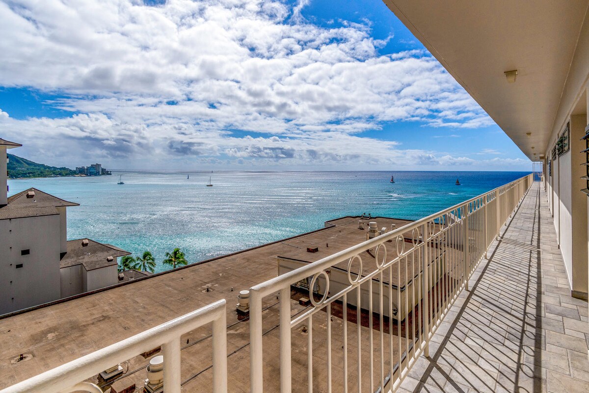 A wide balcony extends along the building, providing views of the ocean featuring multiple boats. The water reflects light, creating a shimmering effect. The sky is dotted with clouds, enhancing the serene atmosphere of the beachside setting.