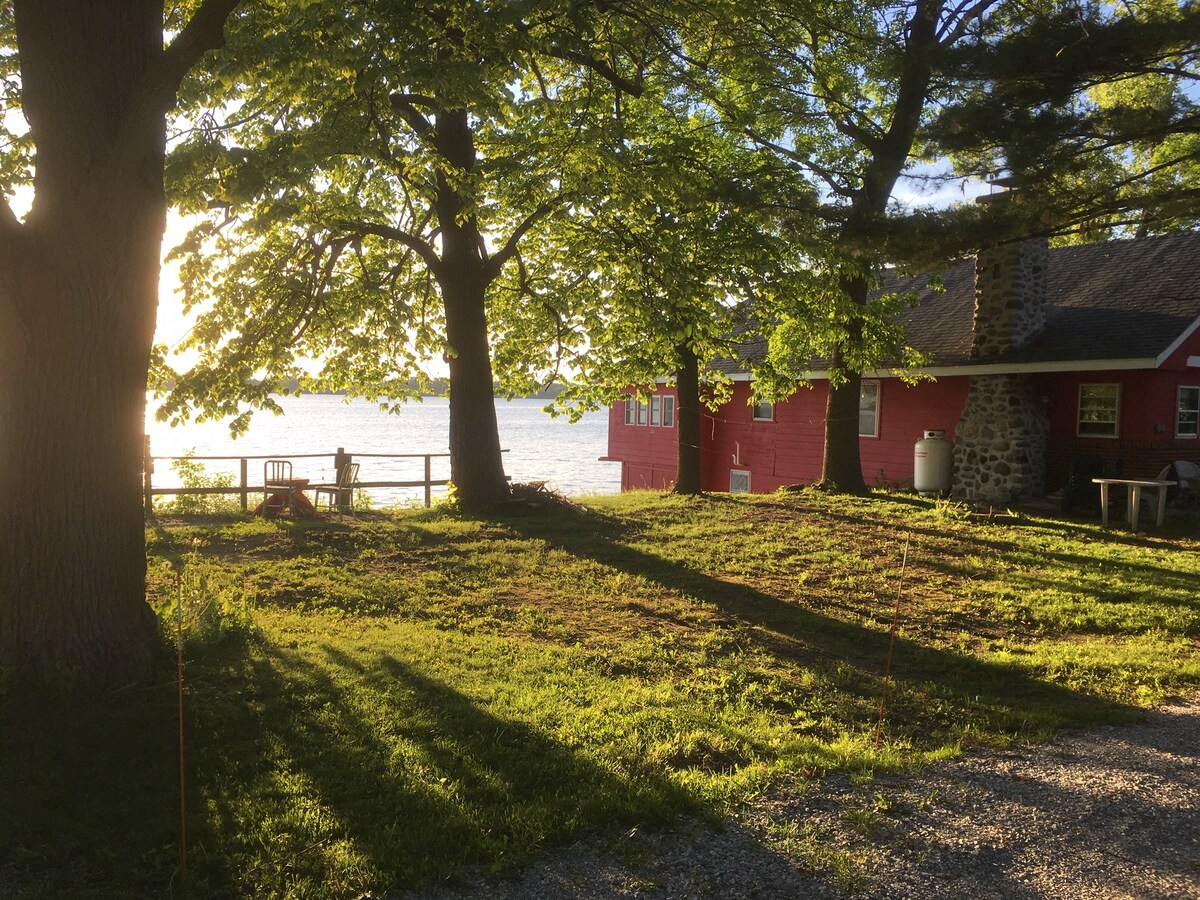 A red rustic cabin stands beside Lake Champlain, framed by large trees that cast long shadows on the grassy lawn. The sun sets in the background, creating a warm glow over the water and enhancing the calm atmosphere of the outdoor space.