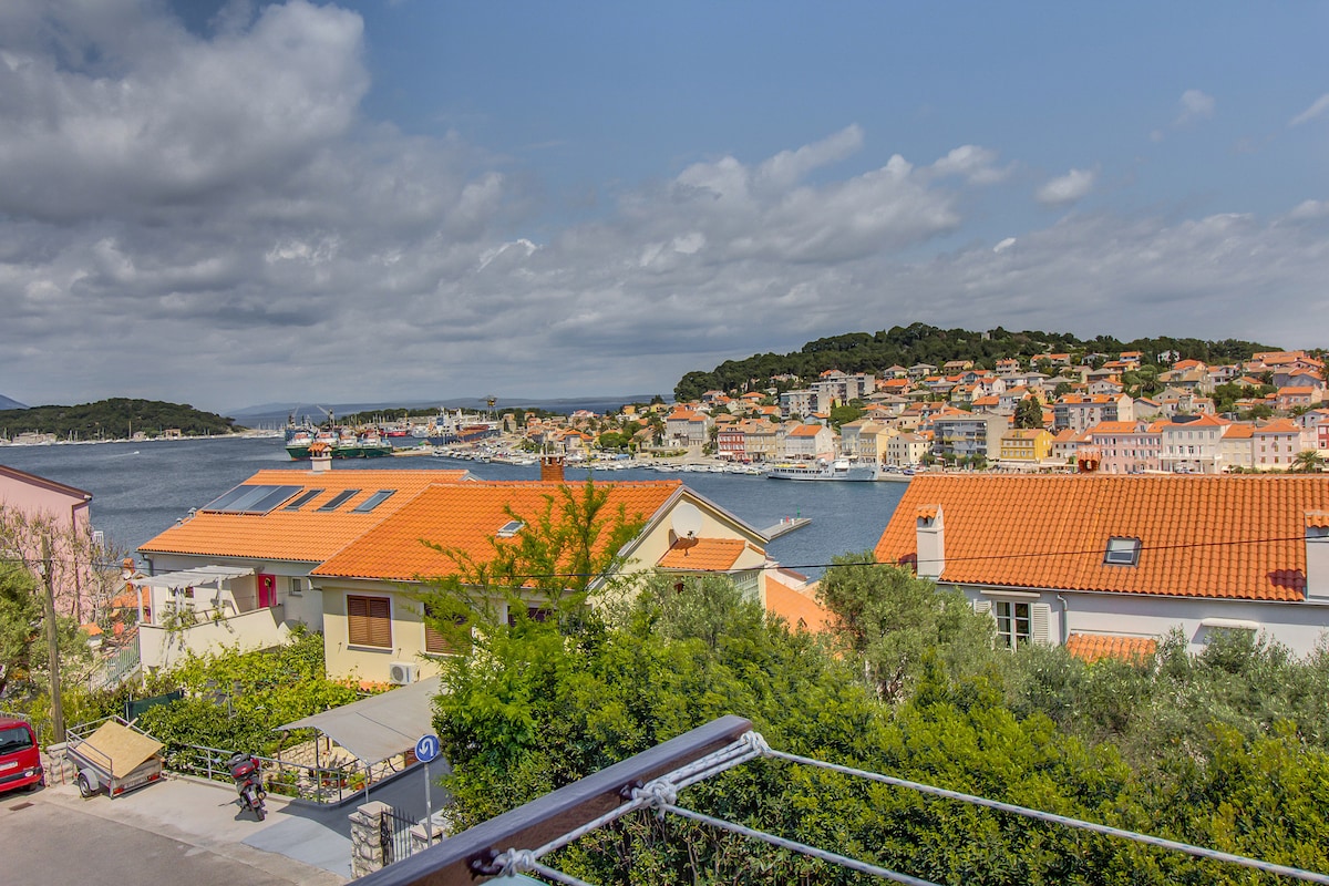 A panoramic view of a coastal town is presented, showcasing colorful rooftops under a partly cloudy sky. The shimmering water reflects the sunlight, while hills and greenery surround the area. Various buildings and boats are visible along the waterfront, creating a lively scene.